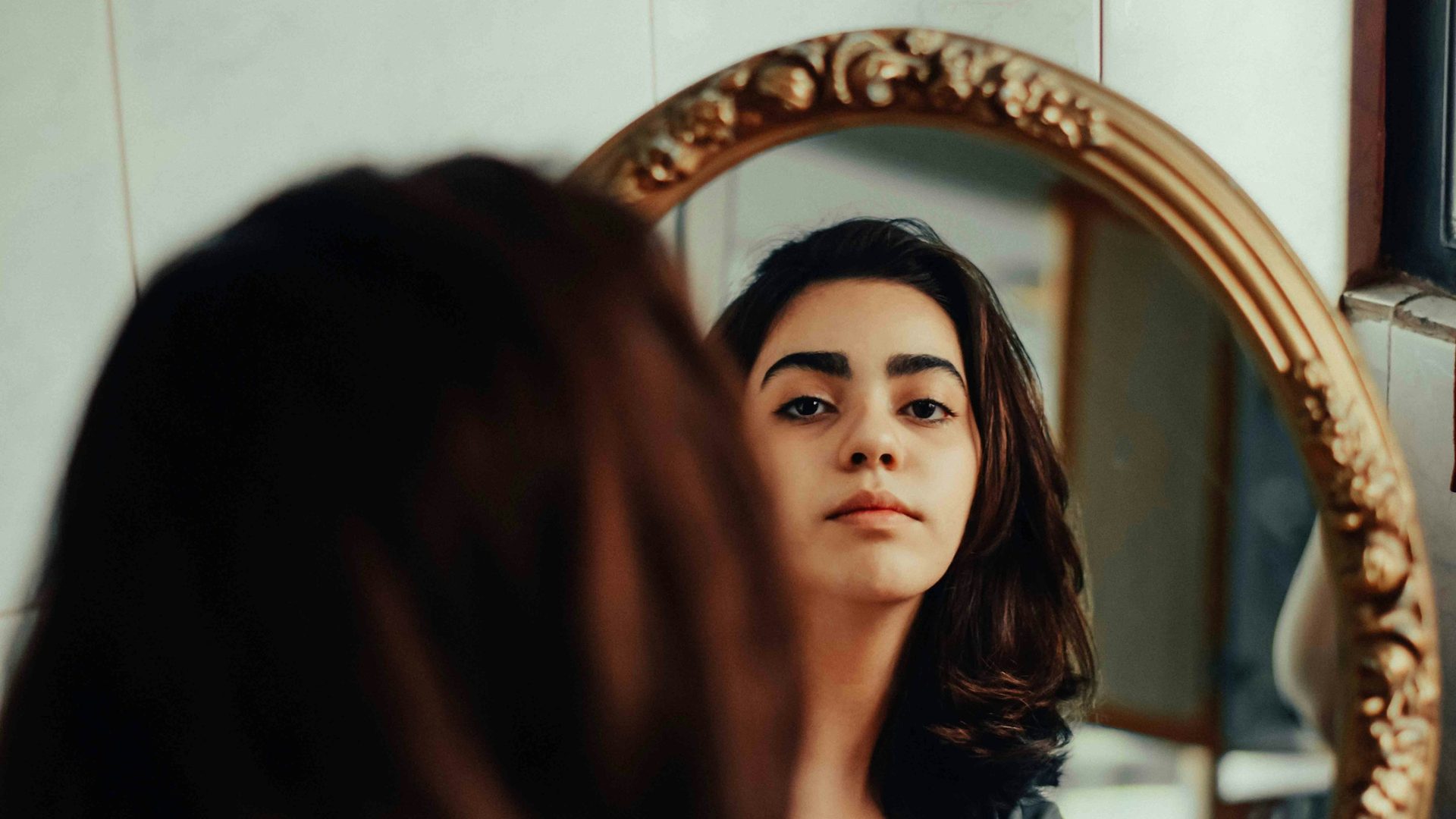 Una giovane donna dai capelli scuri si guarda in uno specchio ovale con cornice dorata